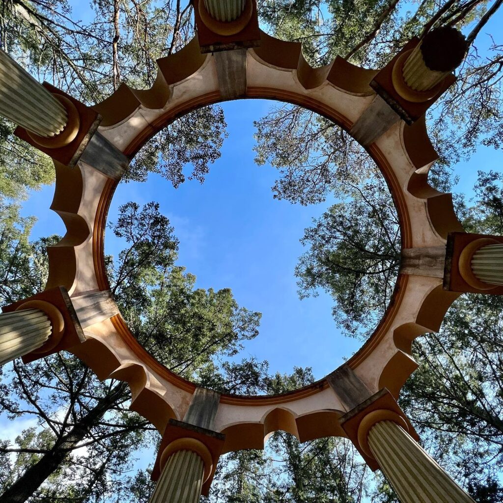 A unique wooden gazebo with a distinctive circular opening framed by arched columns against a clear blue sky.