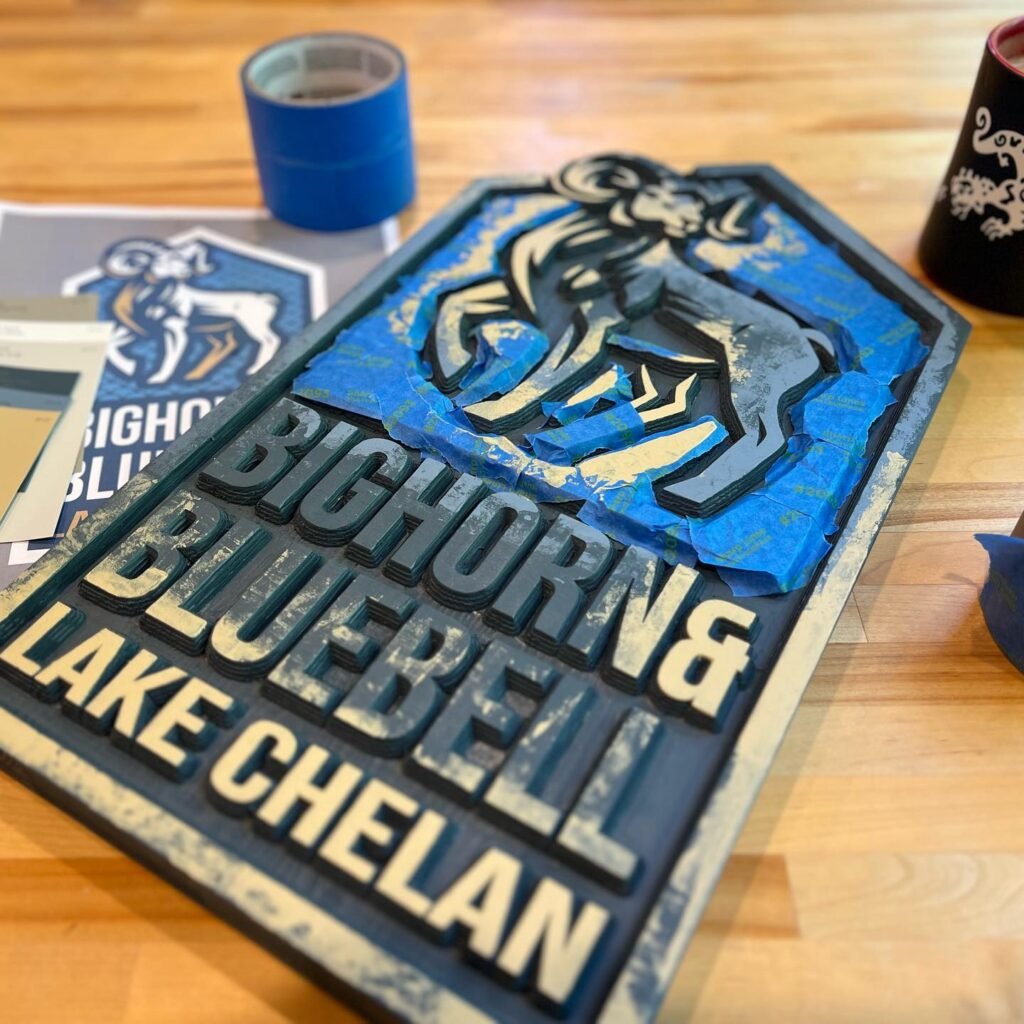 A close-up of a hand-painted sign for Bighorn & Bluebell Lake Chelan, featuring a blue goat logo, a small logo sticker, and crafting supplies on a wooden surface.