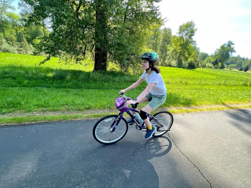 A child rides a purple bicycle on a paved path with vibrant green fields and trees in the background. The scene captures a sunny day in a rural foothills setting.