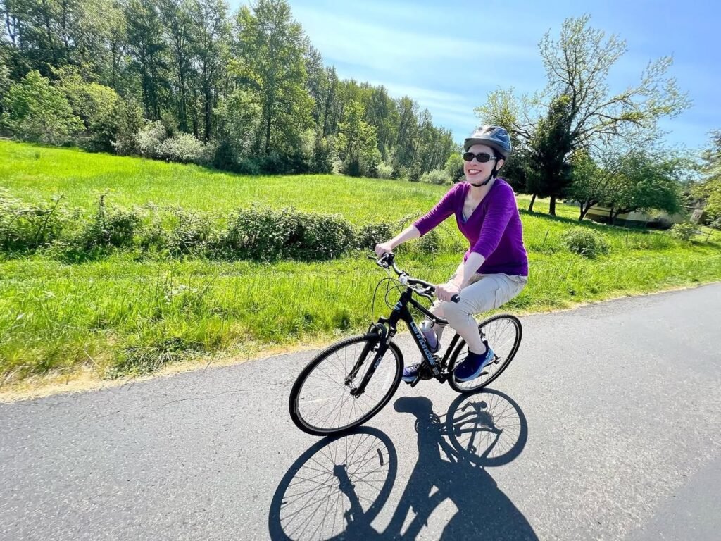 A person rides a bicycle on a paved path surrounded by lush green meadows and tall trees.