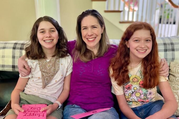 Three smiling people sitting together on a plaid sofa, one holding a pink card. The group appears happy and relaxed in a home setting.