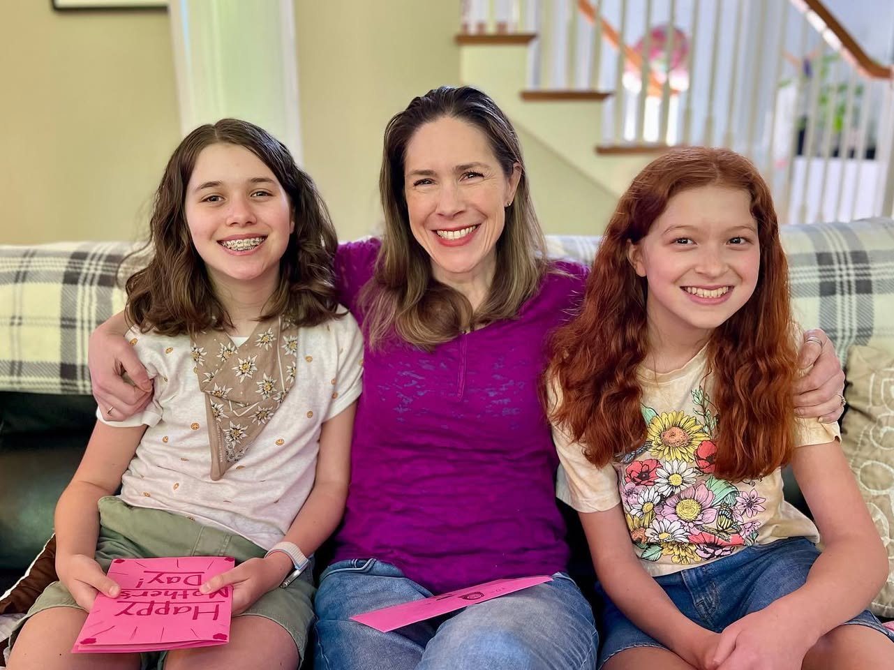 Mother’s Day Bike Ride Three smiling people sitting together on a plaid sofa, one holding a pink card. The group appears happy and relaxed in a home setting.