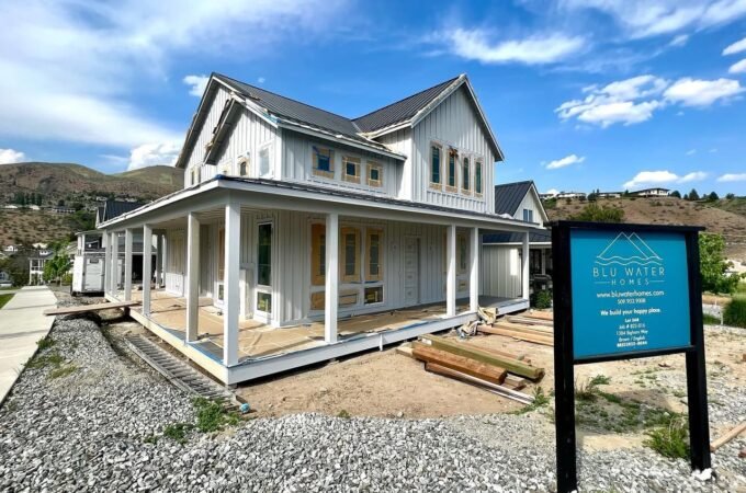 A white colonial house with a front porch set against a clear blue sky and rolling landscape. The property features a gravel foreground and a blue sign nearby.