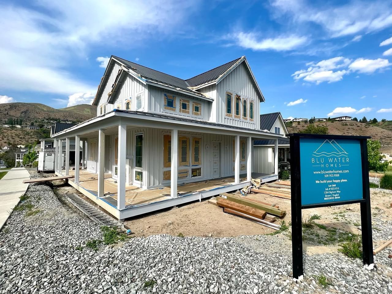 Lake House Progress A white colonial house with a front porch set against a clear blue sky and rolling landscape. The property features a gravel foreground and a blue sign nearby.