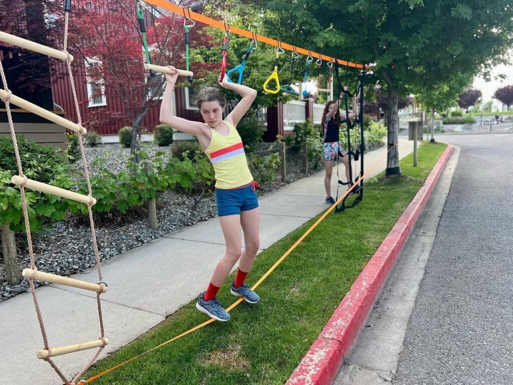 A young person balances on a slackline attached to an orange high rope in a residential backyard. Another individual is visible near a wall that flips in the background setup.