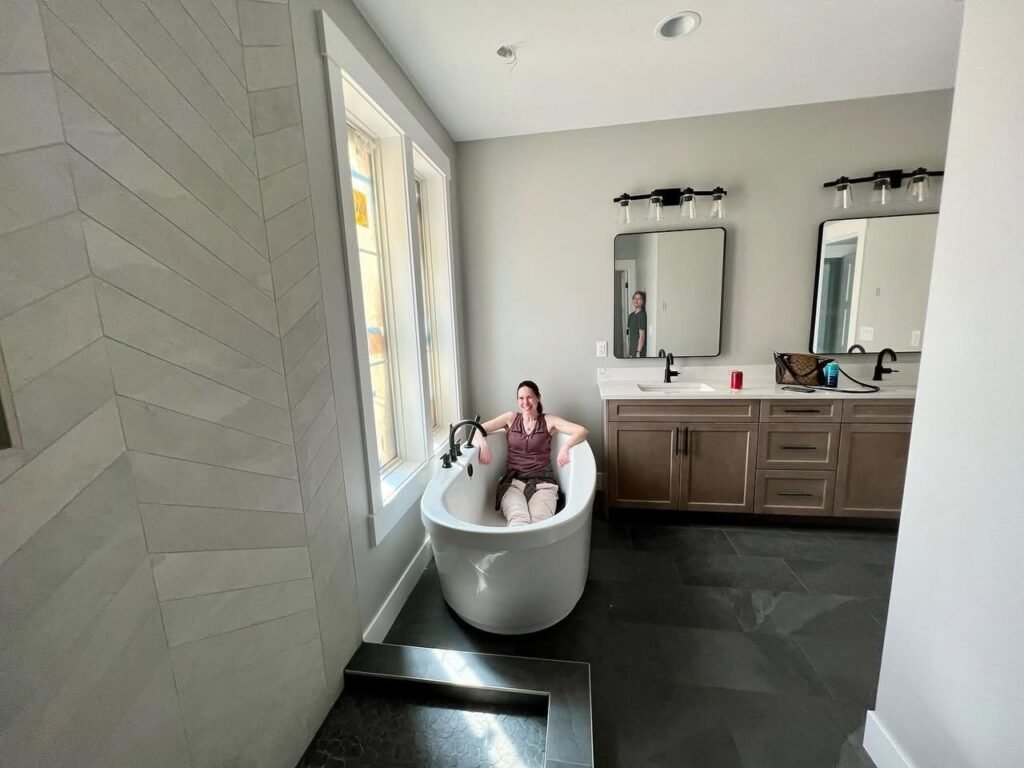 A person relaxes in a white freestanding tub in a modern bathroom with herringbone tile, located at johnandkristie.com.