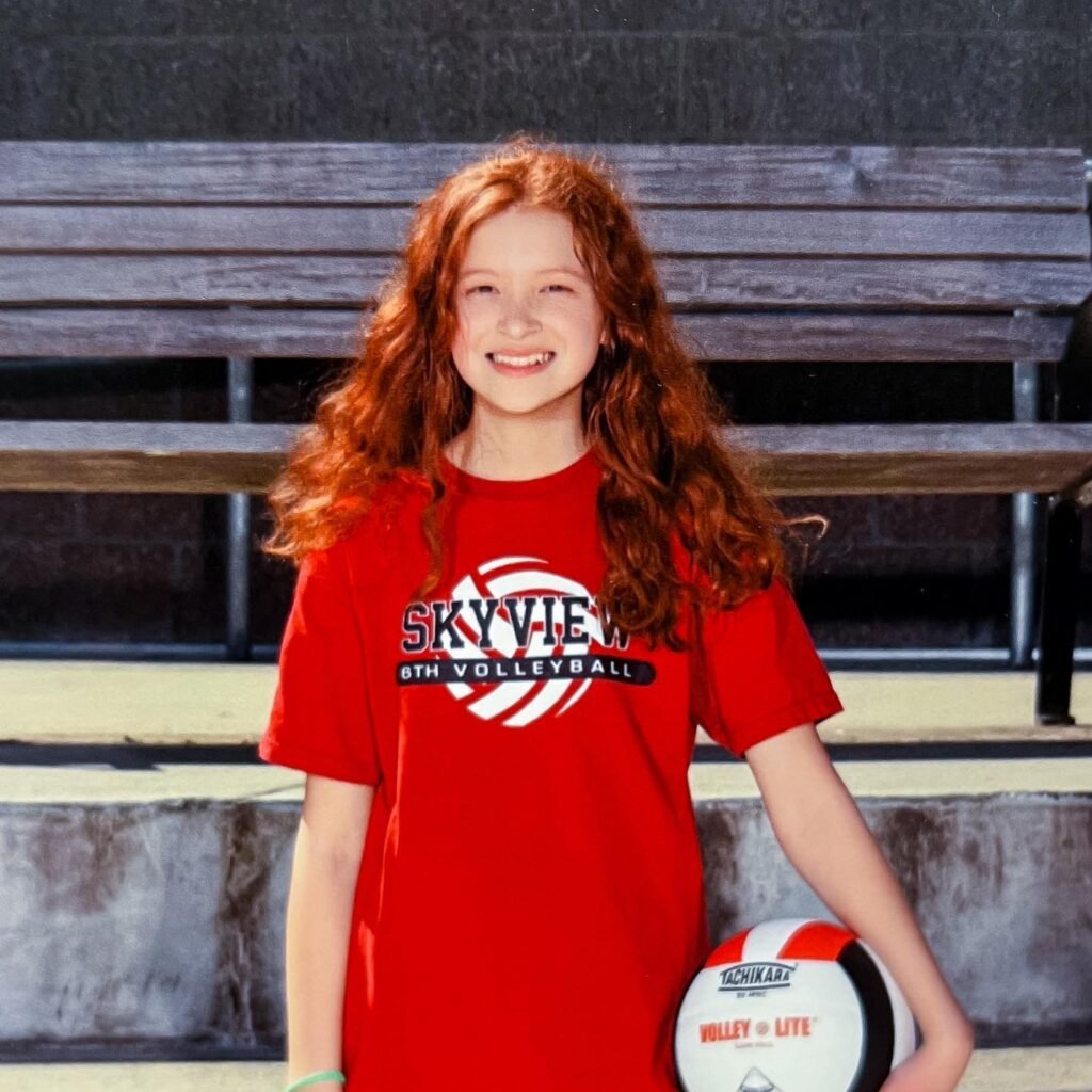 A smiling young athlete with long red hair wearing a red Skyview 6th Volleyball t-shirt and holding a volleyball outdoors.