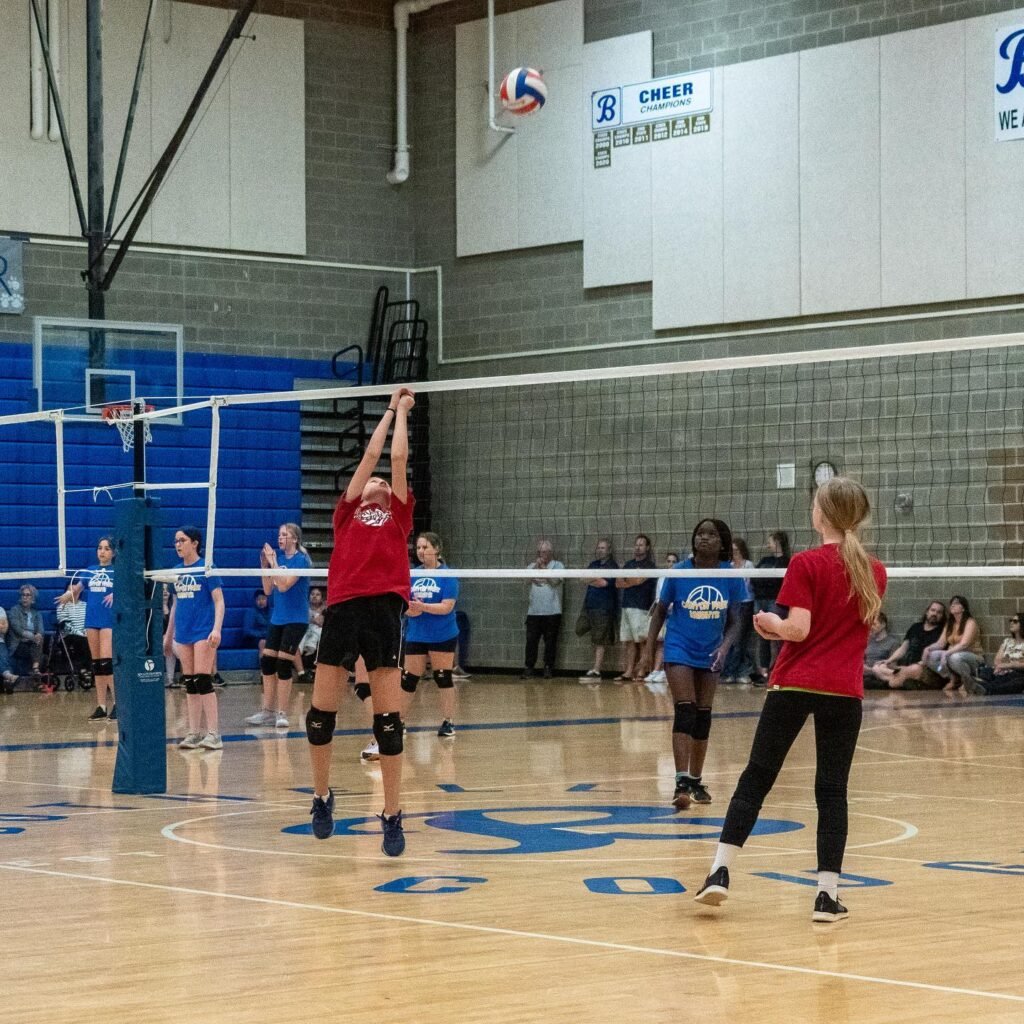 High school volleyball players compete on an indoor court during a match, with a player in a red jersey jumping to spiking the ball over the net.