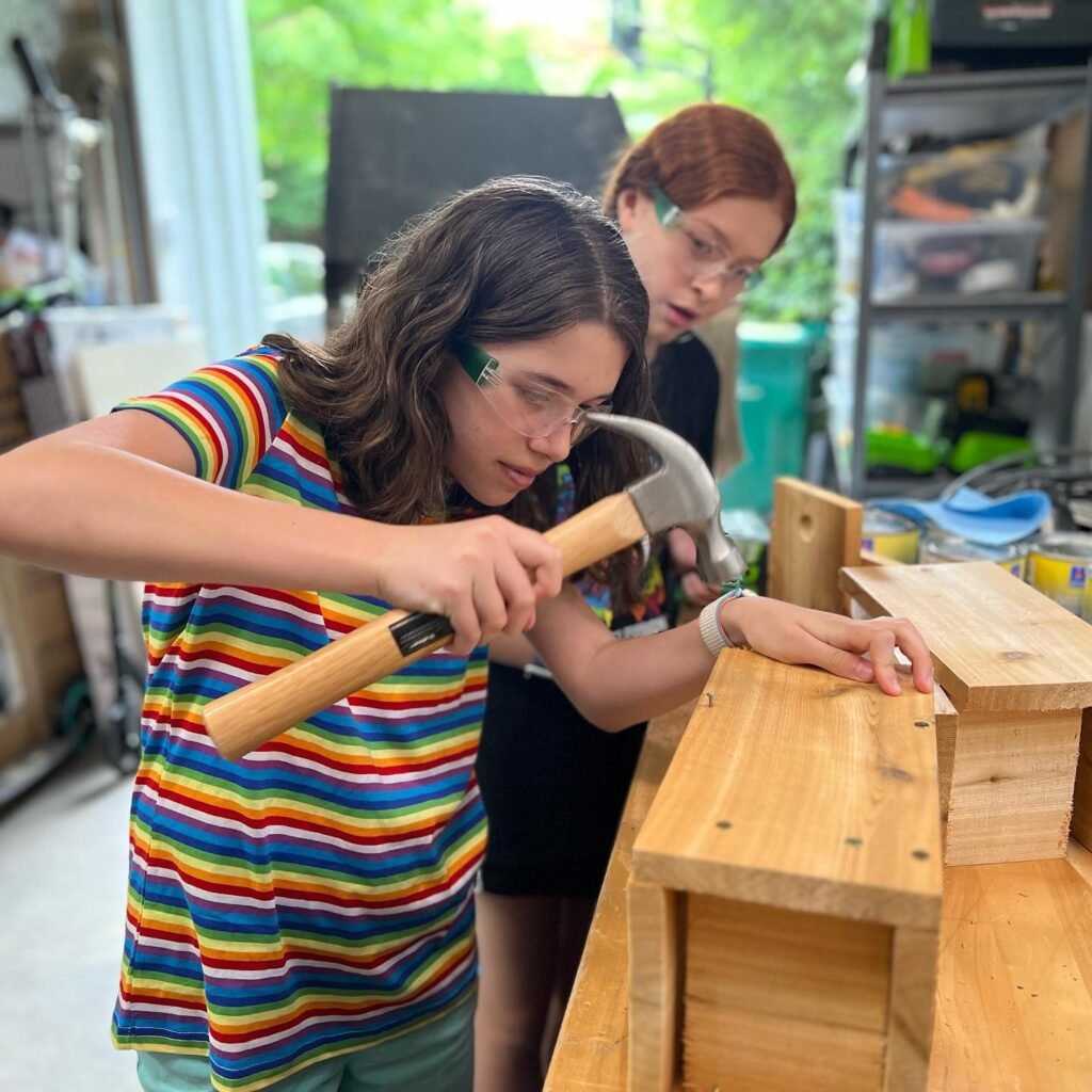 Children wearing safety glasses work with a hammer on a wooden project on a workbench.