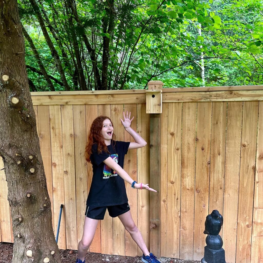 A child poses playfully in a backyard with a lush green cedar tree and a wooden fence.