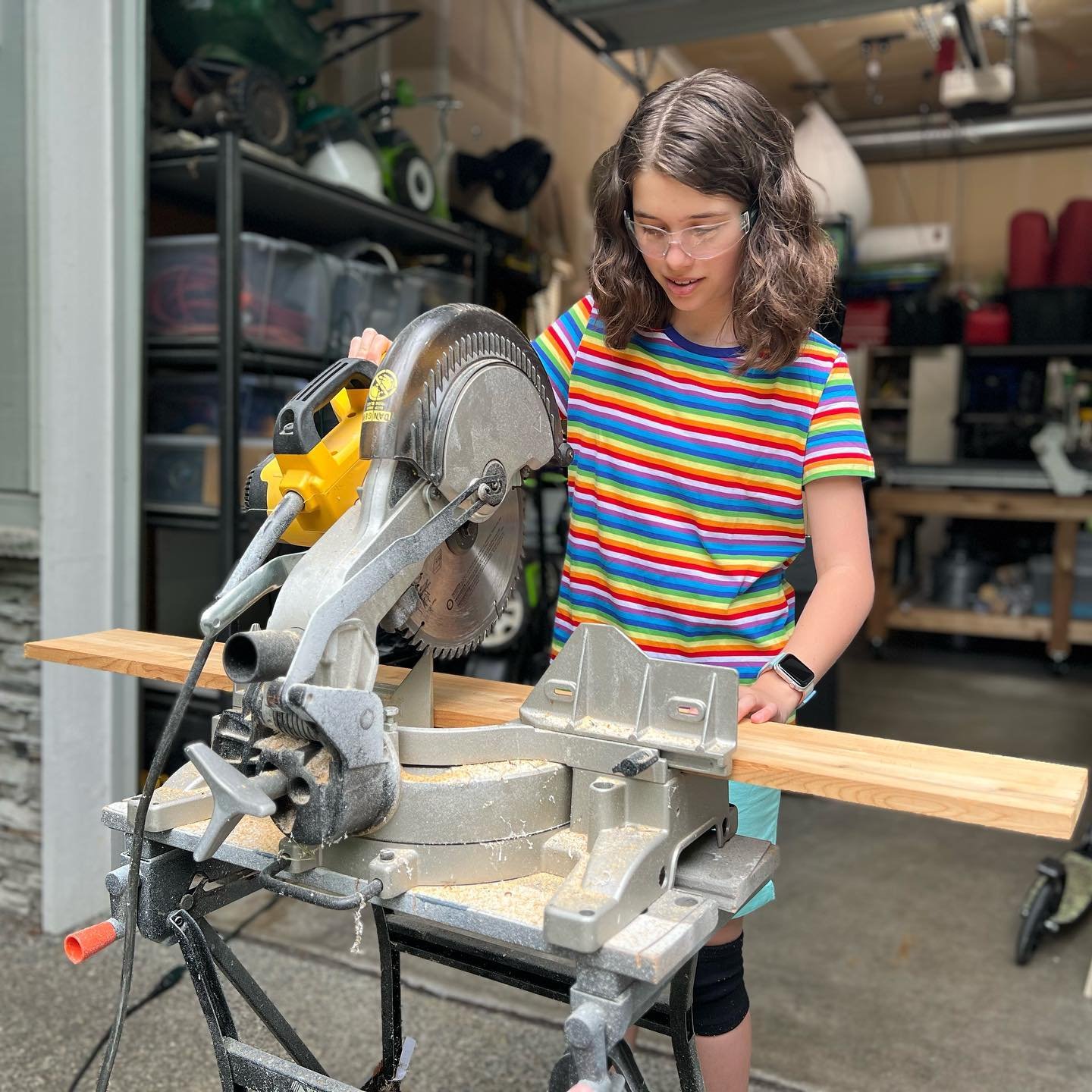 A person wearing safety glasses uses a miter saw in a garage workshop. A close-up shows the blade and the edge of the wooden board being cut.