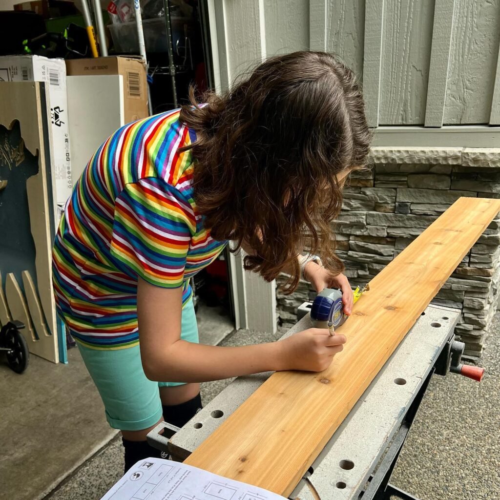 A person in a colorful striped shirt uses a tape measure on a long piece of lumber during a budget-friendly DIY furniture build.