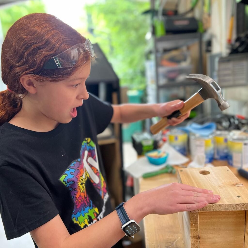A young person wearing safety goggles uses a hammer on a wooden project in a home workshop.