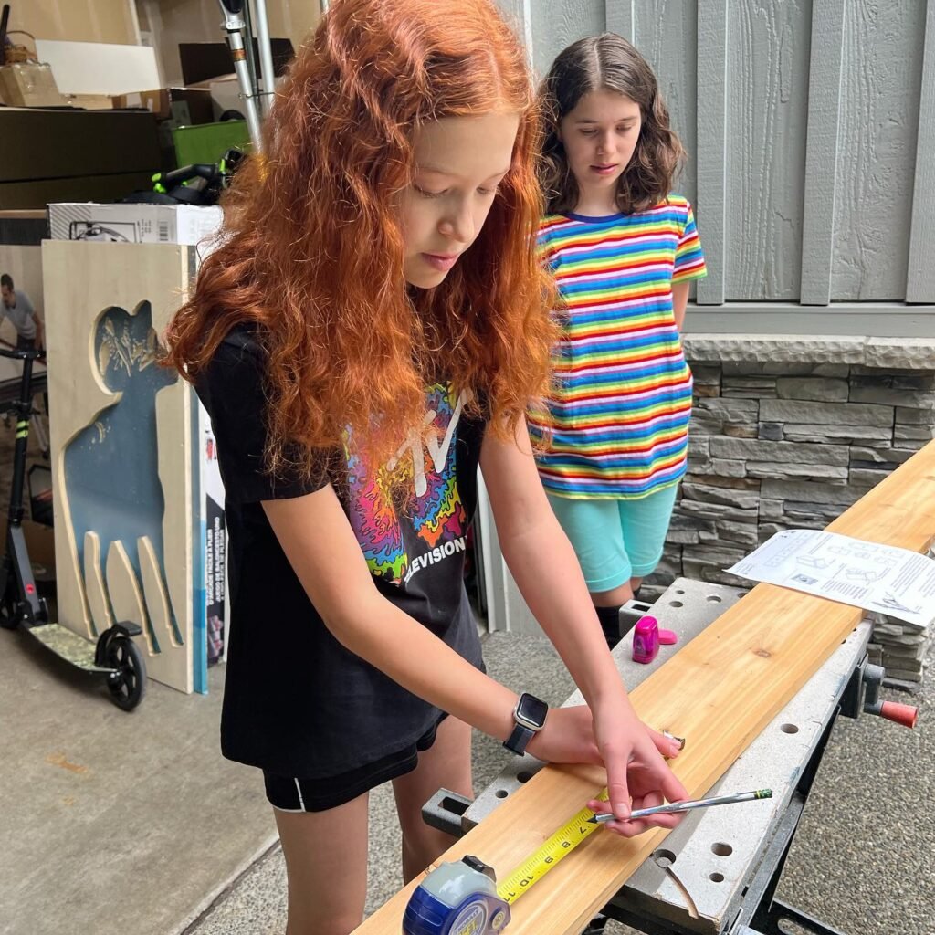 Two girls working on a wooden beam in a sheltered outdoor area, with one person using a tape measure to mark the wood.