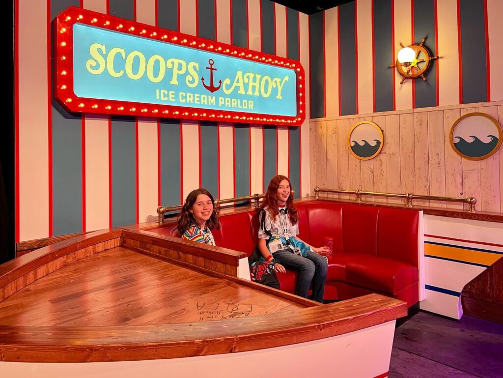 Visitors seated in a nautical-themed red bench under a striped wall at the Scoops Ahoy ice cream stand. A playful pirate flagship interior features bold signage and whimsical design.