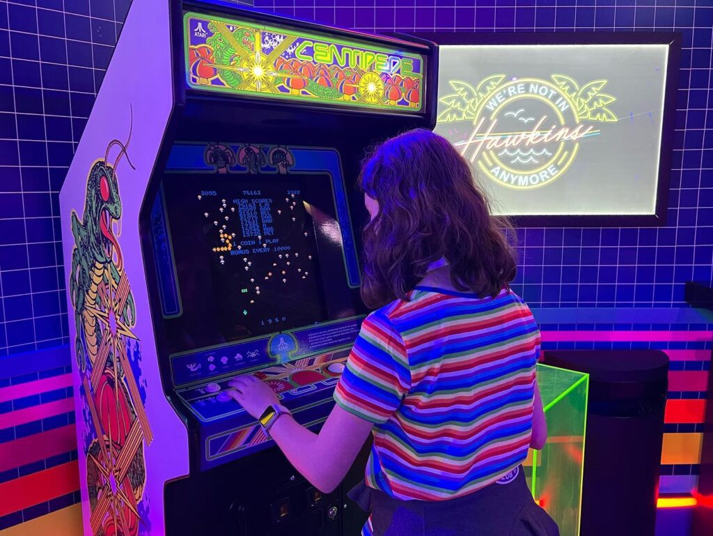 A person plays a colorful Genesis Arcade Warriors machine under vibrant neon lighting.