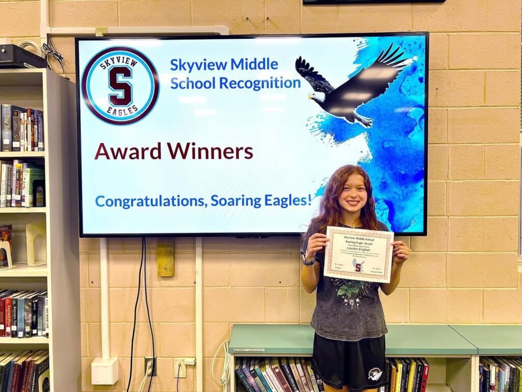 A student proudly displays a certificate in a library in front of a Skyview Middle School award recognition screen.