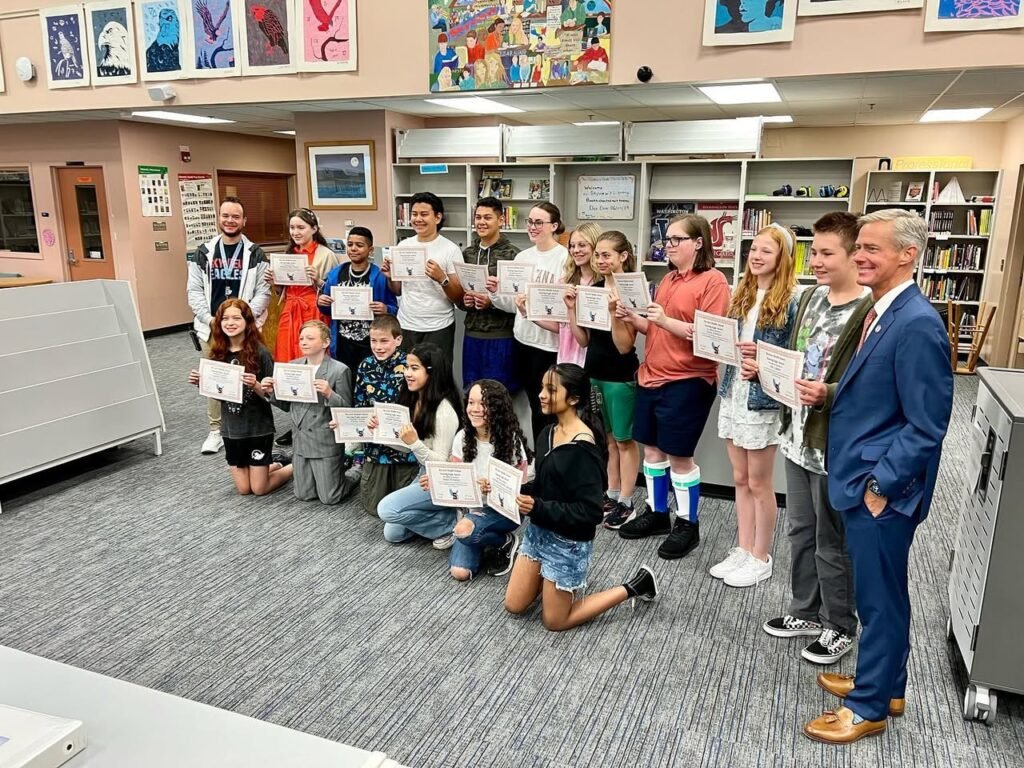 A group photo of students and a man in a bookfilled room, holding certificates and smiling for the camera.