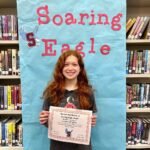 A smiling person holding a certificate in front of a banner reading 'Soaring Eagle' in a library.