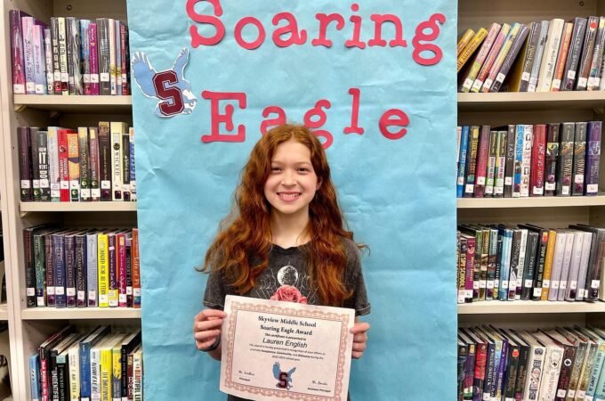 A smiling person holding a certificate in front of a banner reading 'Soaring Eagle' in a library.