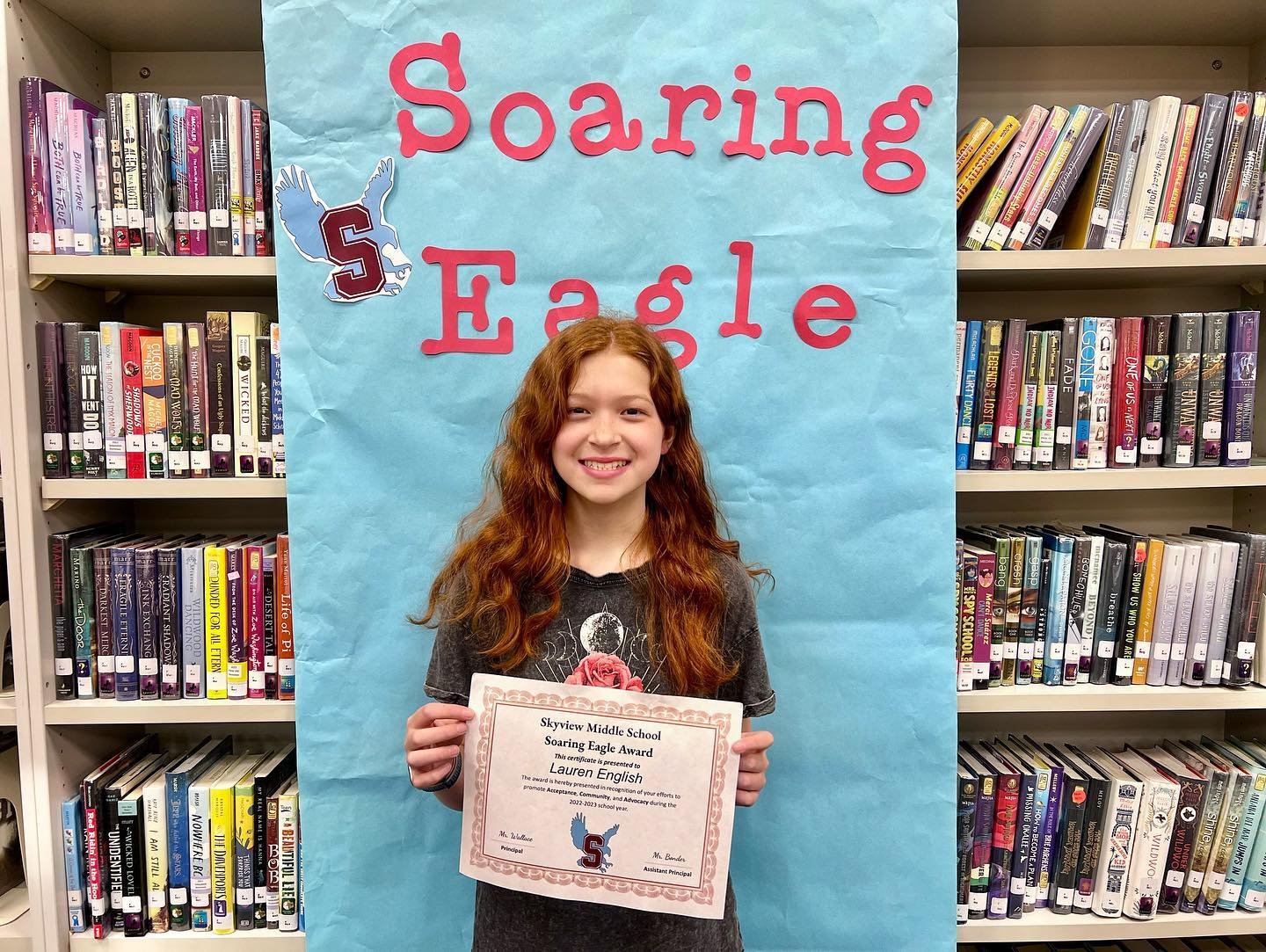 Soaring Eagle Award A smiling person holding a certificate in front of a banner reading 'Soaring Eagle' in a library.