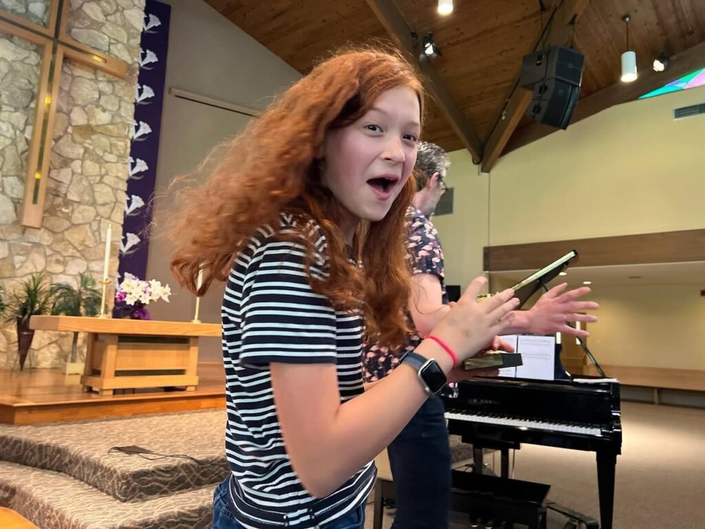 A person with red hair singing while holding a smartphone in a church or chapel setting.