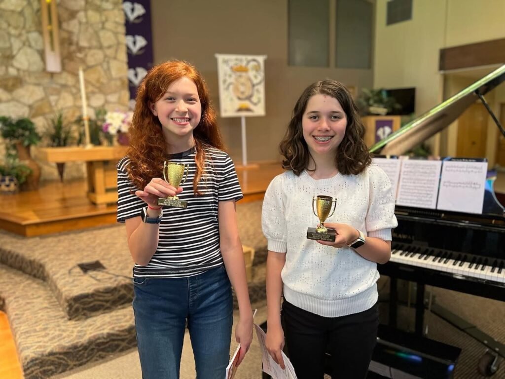 Two smiling individuals standing together holding small trophies next to a grand piano inside a church.
