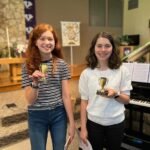 Two smiling individuals standing together holding small trophies next to a grand piano inside a church.