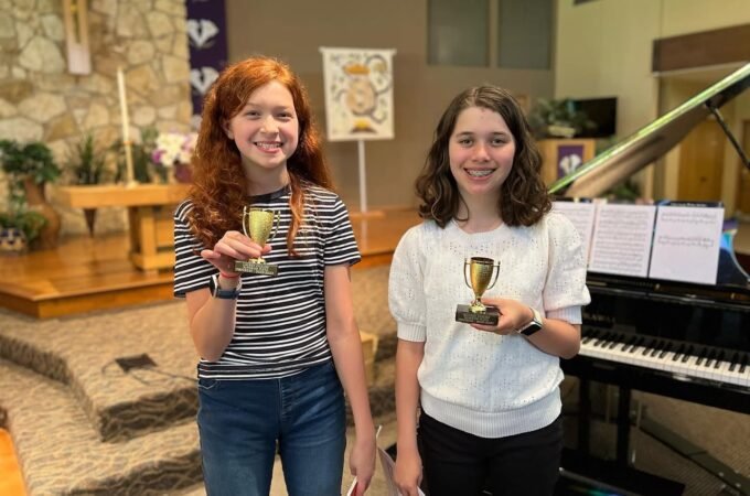 Two smiling individuals standing together holding small trophies next to a grand piano inside a church.