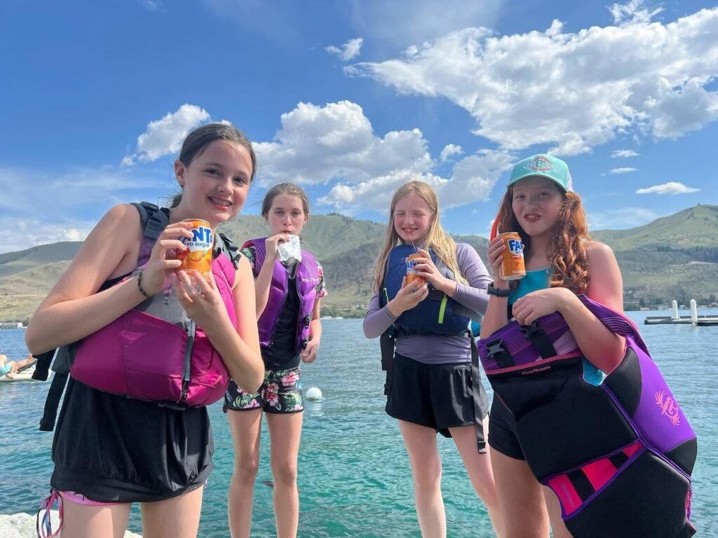 Four girls wearing life jackets smiling with water bottles on a boat, enjoying a sunny day with mountains in the background.