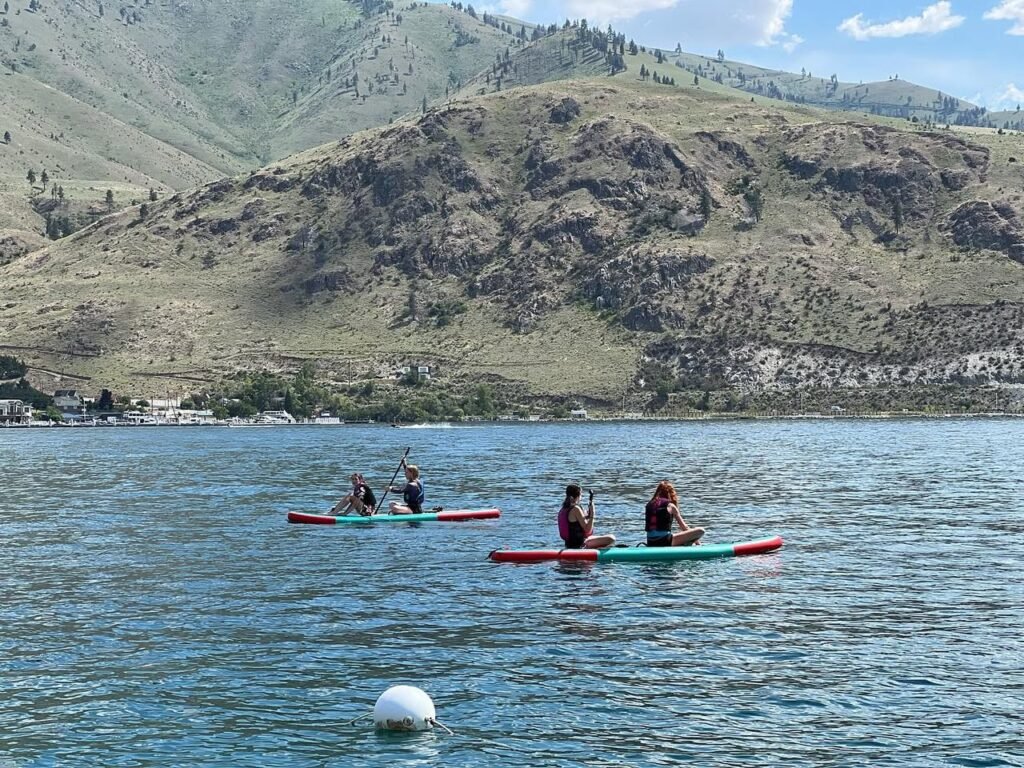 People in a tandem kayak paddling on a calm lake surrounded by rugged mountain terrain.