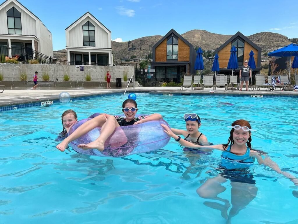 Friends enjoying a sunny day in a resort pool with stylish A-frame cabins and desert hills in the background.