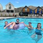 Friends enjoying a sunny day in a resort pool with stylish A-frame cabins and desert hills in the background.