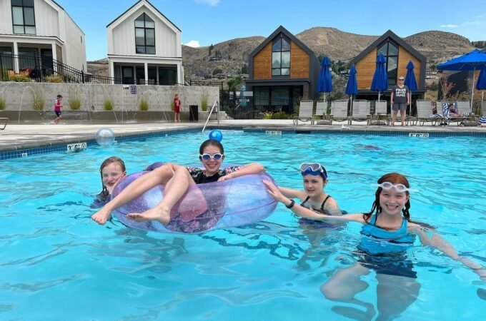 Friends enjoying a sunny day in a resort pool with stylish A-frame cabins and desert hills in the background.