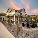 A stunning coastal retreat with a wraparound porch, set against a vibrant sunset sky and rolling hills. The home features modern design elements and a well-maintained landscape.