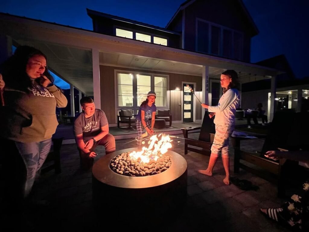 A group of people gathered around a fire pit outside a modern house at dusk, enjoying the warm glow.