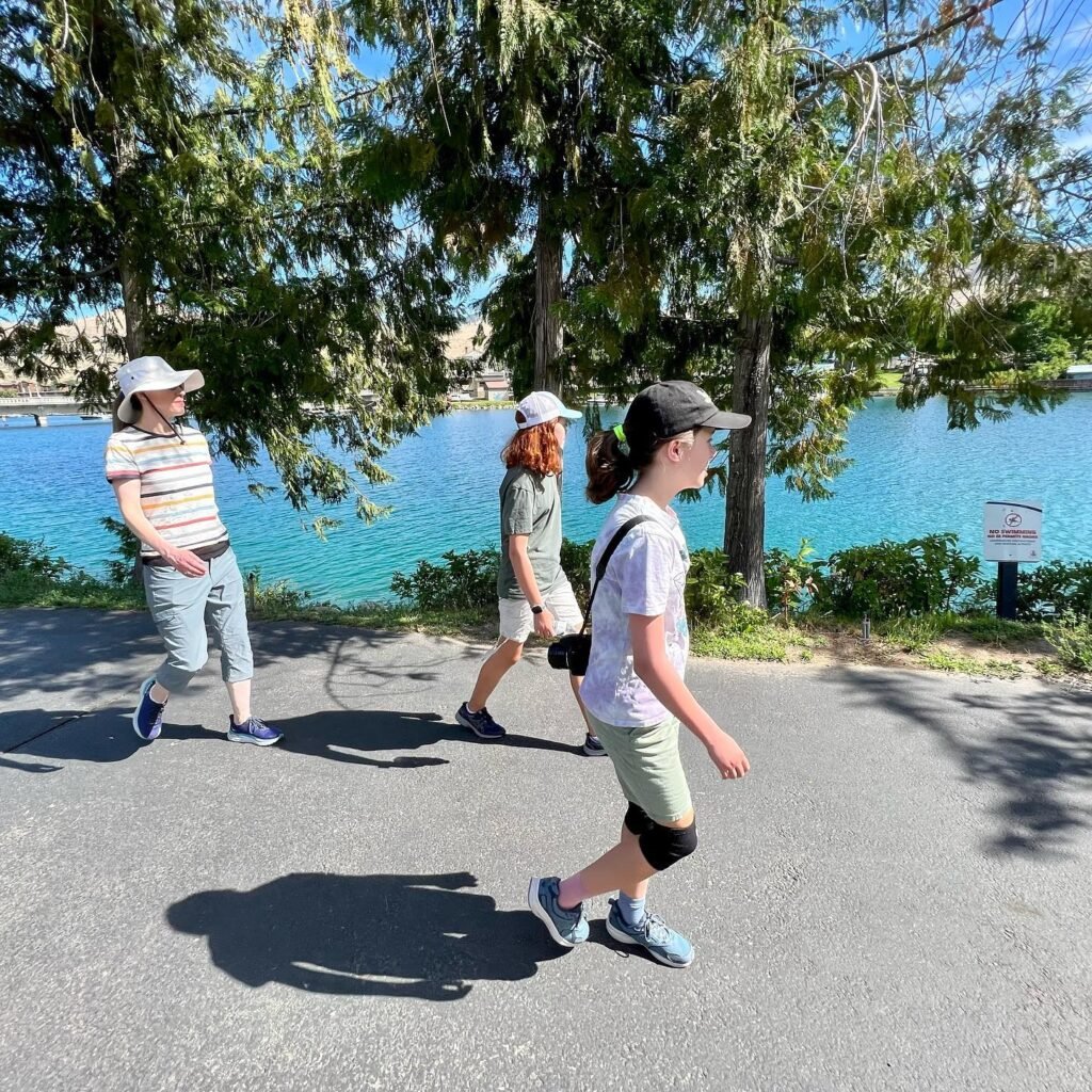 A family enjoys a sunny day walking on a paved path near a bright blue lake. Tall green trees provide shade over the scenic water.