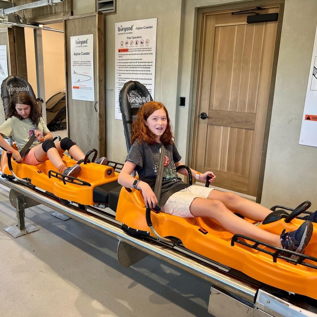 Two children sitting in a bungee jump simulator with orange restraints. The scene takes place in an indoor facility with information posters on the wall.