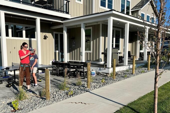 Exterior view of a modern home with covered porches, gravel landscaping, and sidewalk. A person in red holding a child stands on the porch next to picnic tables.