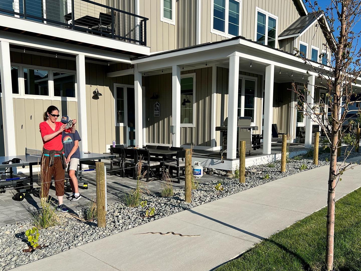 Snake Charmer Exterior view of a modern home with covered porches, gravel landscaping, and sidewalk. A person in red holding a child stands on the porch next to picnic tables.
