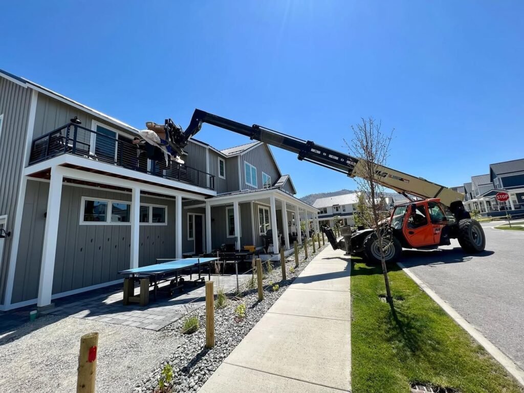 A red tractor equipped with a ball lifter attachment is parked in front of a two-story building with a covered porch and a gravel area.