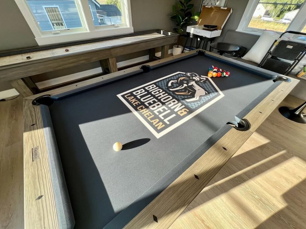 A sunlit photo of a custom pool table on a covered deck featuring a Big Horns Bluebell logo, with billiard balls and a cue resting on the felt.