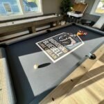 Snake Charmer A sunlit photo of a custom pool table on a covered deck featuring a Big Horns Bluebell logo, with billiard balls and a cue resting on the felt.