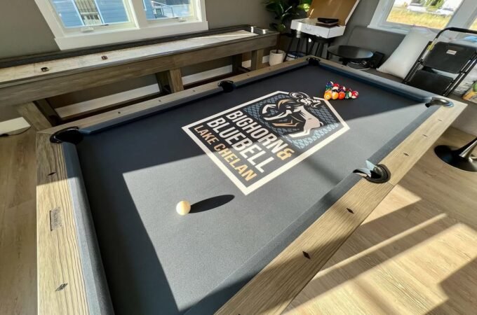 A sunlit photo of a custom pool table on a covered deck featuring a Big Horns Bluebell logo, with billiard balls and a cue resting on the felt.