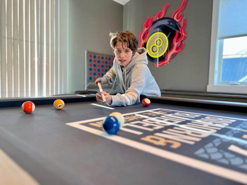 A person leans over a home billiards table to make a shot. The scene features a decorative flaming 8-ball on the wall and several colorful balls on the felt.