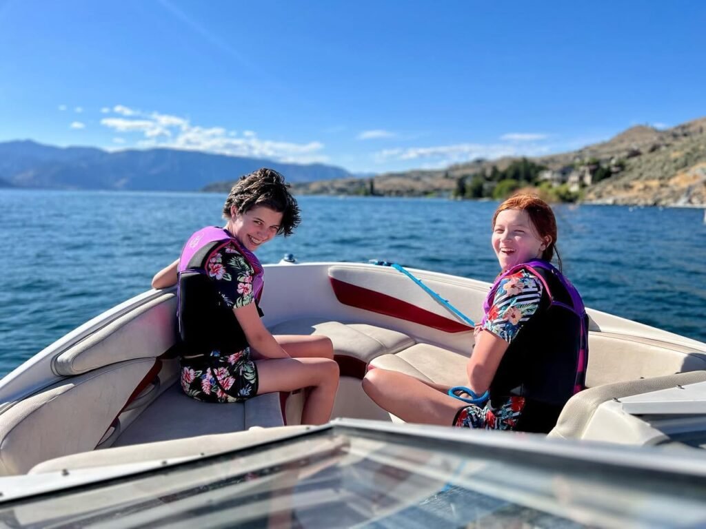 Two young people wearing life jackets sit on a boat, surrounded by clear blue water and scenic mountain views.