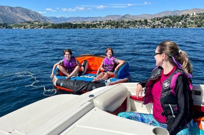 Several people wearing life jackets are swimming and using a raft near a boat on a bright, sunny day in the Okanagan Valley.