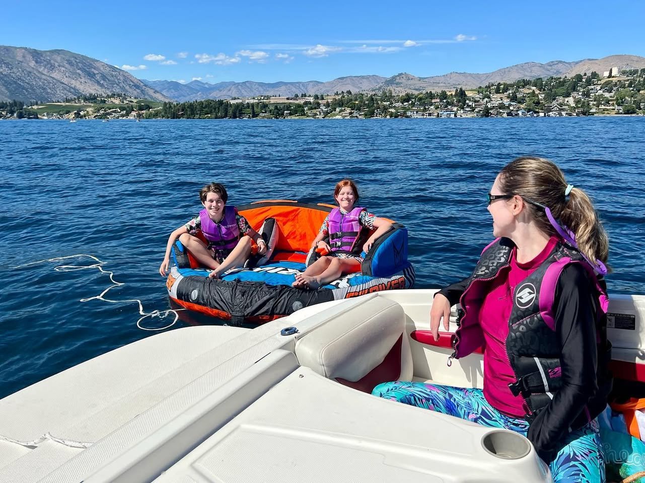Several people wearing life jackets are swimming and using a raft near a boat on a bright, sunny day in the Okanagan Valley.