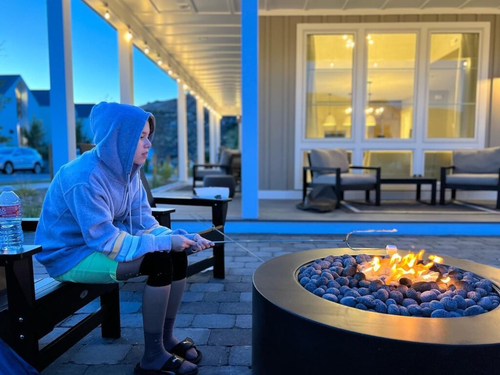 A person in a hoodie enjoys a campfire by a pool in the evening. A warmly lit house and deck area are visible in the background.
