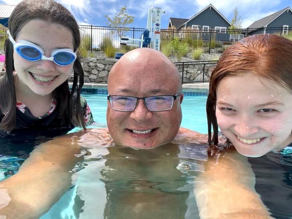 Smiling individuals enjoy a swim in an outdoor pool with a house and bungee tower in the background.